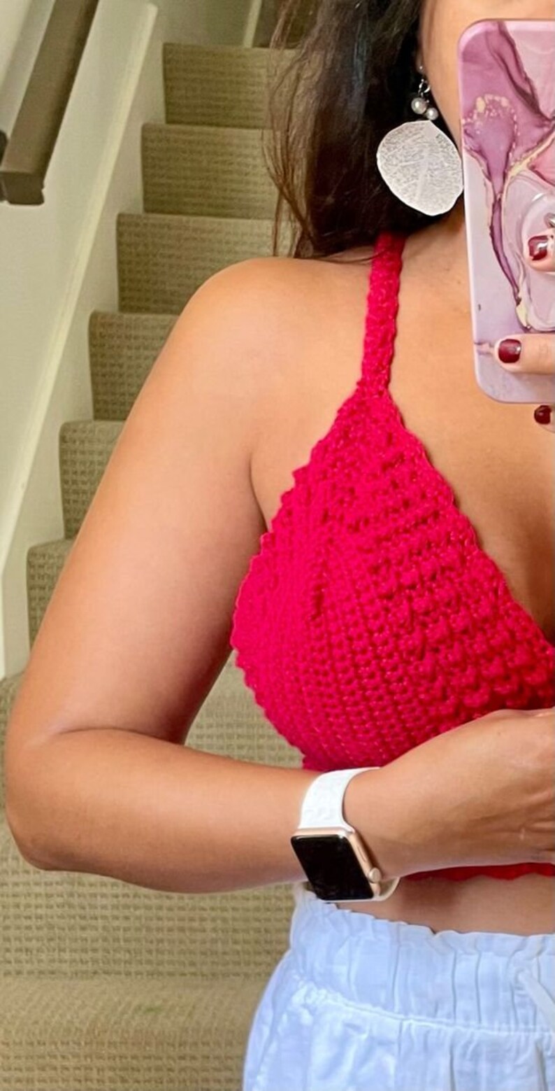 May include: A bright red, handmade crochet halter top. The top is a solid color and has a textured appearance. The straps are thick and the top is a triangle shape. A white watch with a gold case is visible on the wrist.