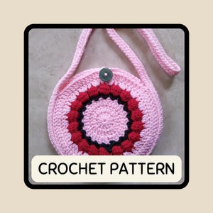 Op de afbeelding: Een roze gehaakte ronde tas met een riem, met een centraal ontwerp van rode en zwarte bobbles. De tas heeft een knoopsluiting en de tekst "CROCHET PATTERN" wordt onderaan weergegeven.
