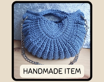 May include: A handmade blue crochet bag with a silver chain strap. The bag has a unique, textured design and a curved shape. The text "HANDMADE ITEM" is displayed in a white rectangle at the bottom of the image.