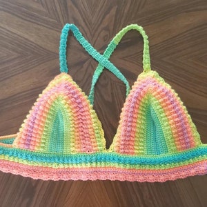 May include: A handmade, colorful crochet top in a rainbow of hues. The top features a triangle shape with straps that cross in the back. The colors include pink, orange, yellow, green, and blue, creating a vibrant and eye-catching design.