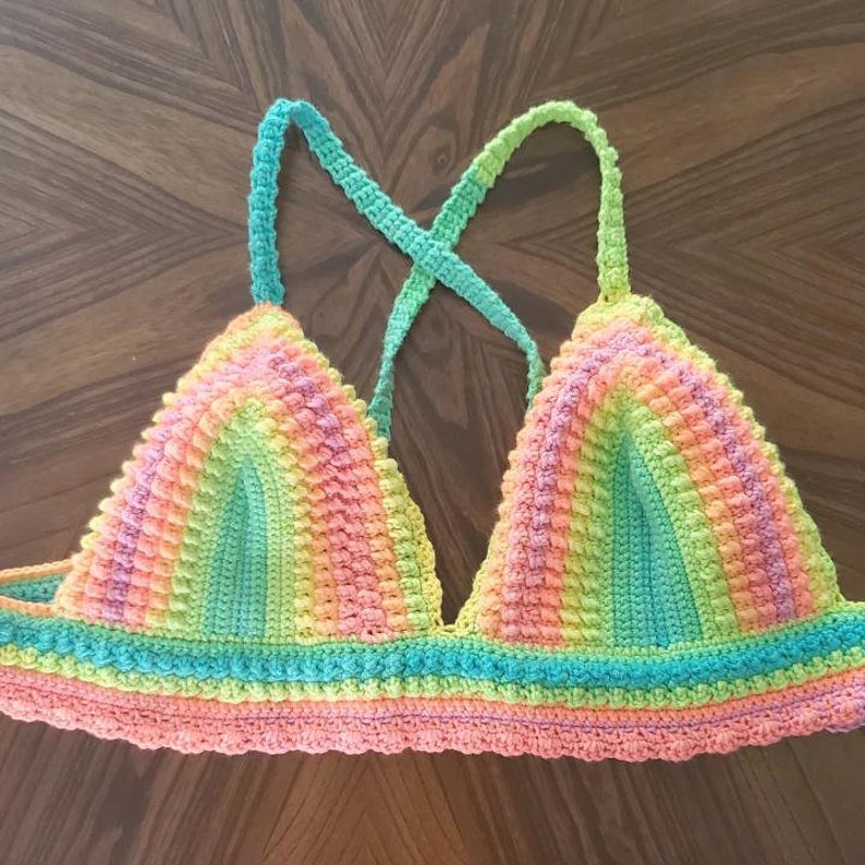 May include: A handmade, colorful crochet top in a rainbow of hues. The top features a triangle shape with straps that cross in the back. The colors include pink, orange, yellow, green, and blue, creating a vibrant and eye-catching design.