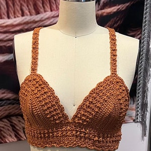 May include: A handmade, rust-colored crochet top with a V-neck design. The top features textured stitching and delicate straps. The garment is displayed on a mannequin, showcasing its intricate details and craftsmanship.