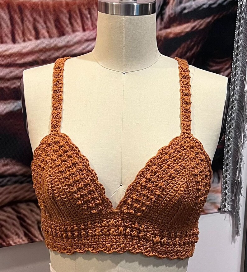 May include: A handmade, rust-colored crochet top with a V-neck design. The top features textured stitching and delicate straps. The garment is displayed on a mannequin, showcasing its intricate details and craftsmanship.