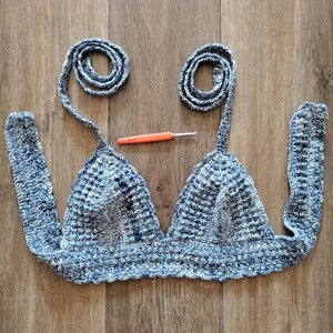 May include: A handmade blue and white crochet top with long straps and a matching crochet hook. The top features a triangle shape with a textured stitch pattern. The straps are rolled into a spiral shape.