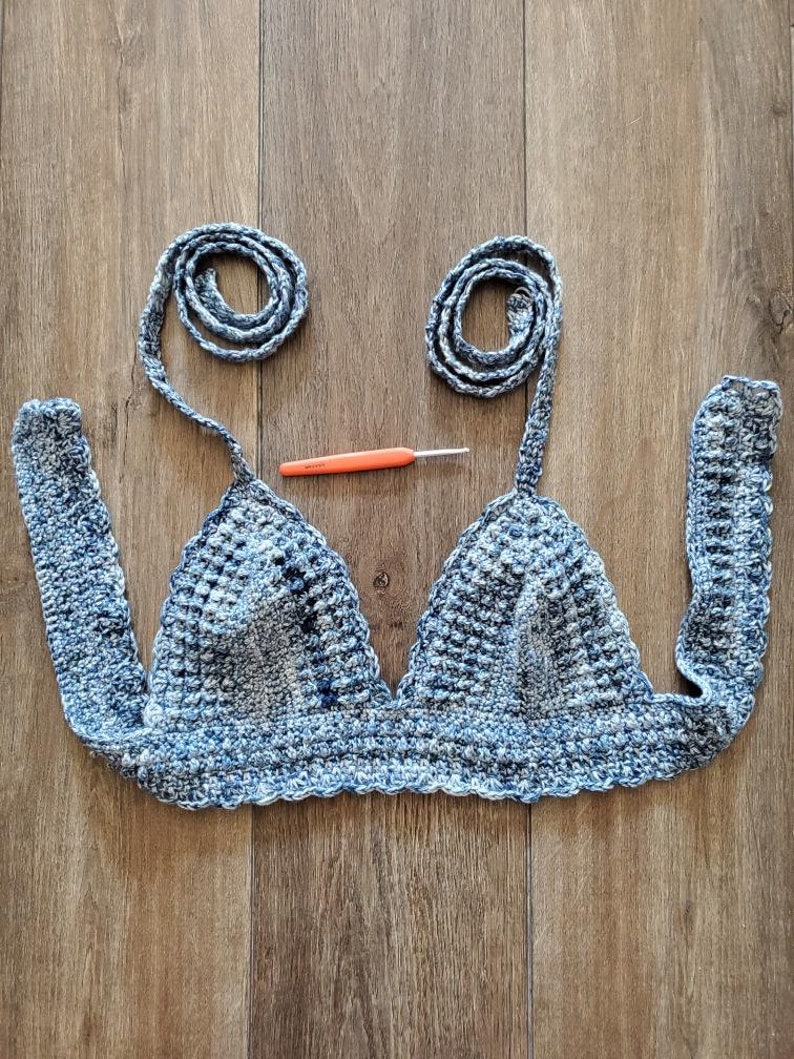 May include: A handmade blue and white crochet top with long straps and a matching crochet hook. The top features a triangle shape with a textured stitch pattern. The straps are rolled into a spiral shape.