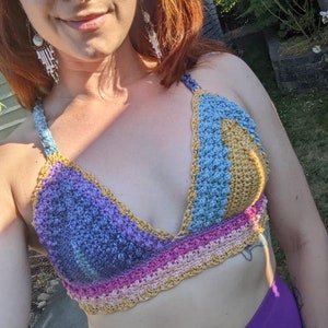 May include: A handmade crochet top in a variety of colors, including blue, purple, pink, and gold. The top has a halter neck design and a textured stitch pattern. The garment is worn with purple pants.