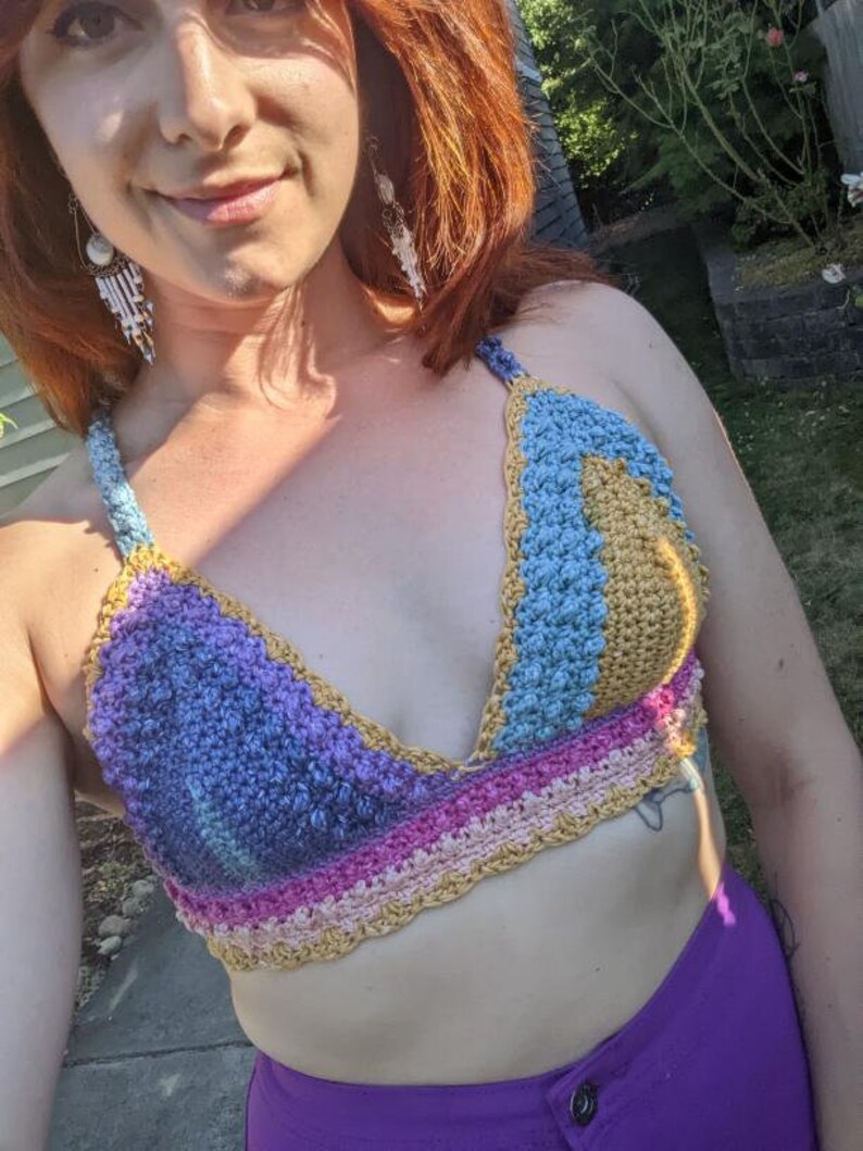 May include: A handmade crochet top in a variety of colors, including blue, purple, pink, and gold. The top has a halter neck design and a textured stitch pattern. The garment is worn with purple pants.