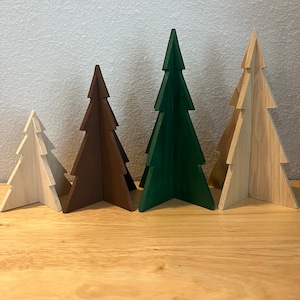 May include: Four wooden Christmas tree ornaments in different colors: white, brown, green, and natural wood. The trees are standing on a wooden surface.