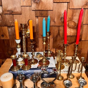 Peut inclure: Une collection de bougeoirs en laiton vintage avec différents designs, dont un bougeoir en forme de cœur, un bougeoir en forme de trompette et un bougeoir à base ronde. Les bougeoirs sont disposés sur une surface en bois avec un livre et une bougie blanche.