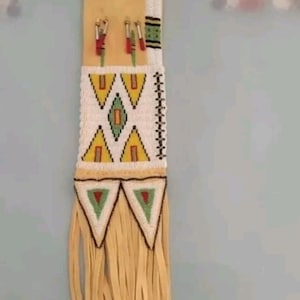 Peut inclure: Une pochette amérindienne en cuir ornée de perles. La pochette est décorée d'un motif géométrique en perles blanches, jaunes, vertes et rouges. La pochette mesure environ 30 cm de long.