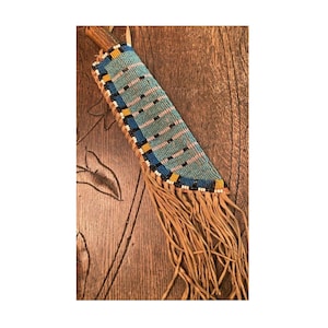 Op de afbeelding: Een kralen schede voor een mes met een franje. De schede is gemaakt van leer en heeft een kralenpatroon in blauw, wit en geel. De franje is van leer en is bruin.