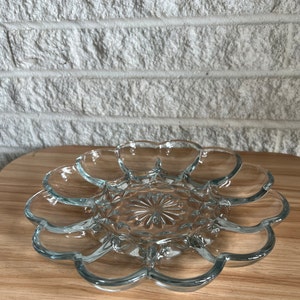 May include: Clear glass serving dish with a flower-like design. The dish has multiple compartments and a central starburst pattern. It sits on a wooden surface, with a white brick wall in the background.