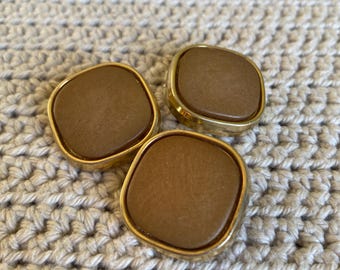 May include: Three brown square buttons with gold-colored metal rims. The buttons have a slightly rounded shape and a smooth, matte finish. The buttons are arranged on a textured, off-white crocheted surface.