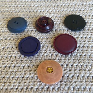 May include: A collection of six buttons in various colors and designs. The buttons include shades of blue, burgundy, and black, with one featuring a gold flower. The buttons are round and have two or four holes for sewing.
