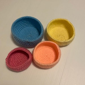 Puede incluir: Cuatro cuencos de ganchillo hechos a mano en tonos azul, amarillo, rosa y melocotón. Los cuencos son redondos con un diseño sencillo y abierto, perfectos para objetos pequeños o uso decorativo. Los cuencos están dispuestos sobre una superficie blanca.