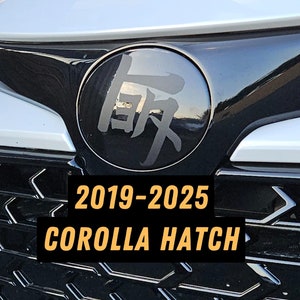 May include: A black and silver car grille with a black circle emblem featuring Japanese characters. The text "2019-2025 COROLLA HATCH" is printed in yellow on the grille.