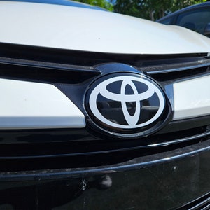 May include: Close-up of a white car's front grille with a black Toyota logo. The logo is a white oval with a stylized 'T' inside.