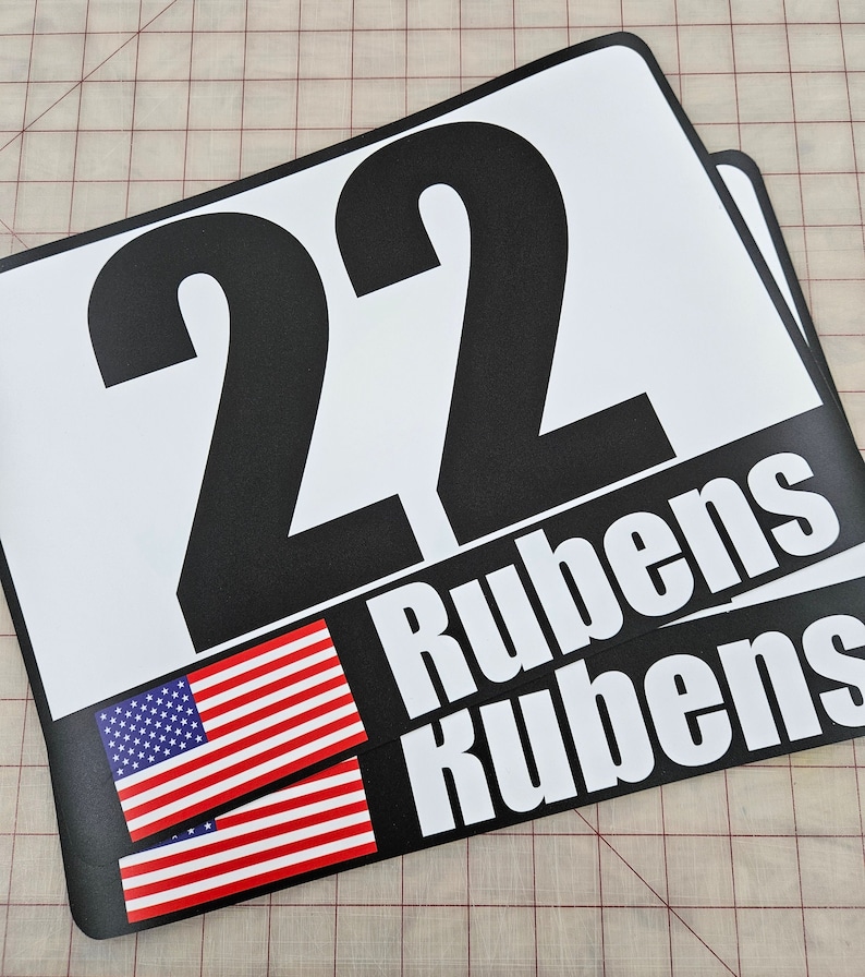 May include: A rectangular white sign featuring the large black number 22 at the top. Below the number, a black horizontal band displays a small American flag icon on the left and the name Rubens printed in white capital letters on the right.