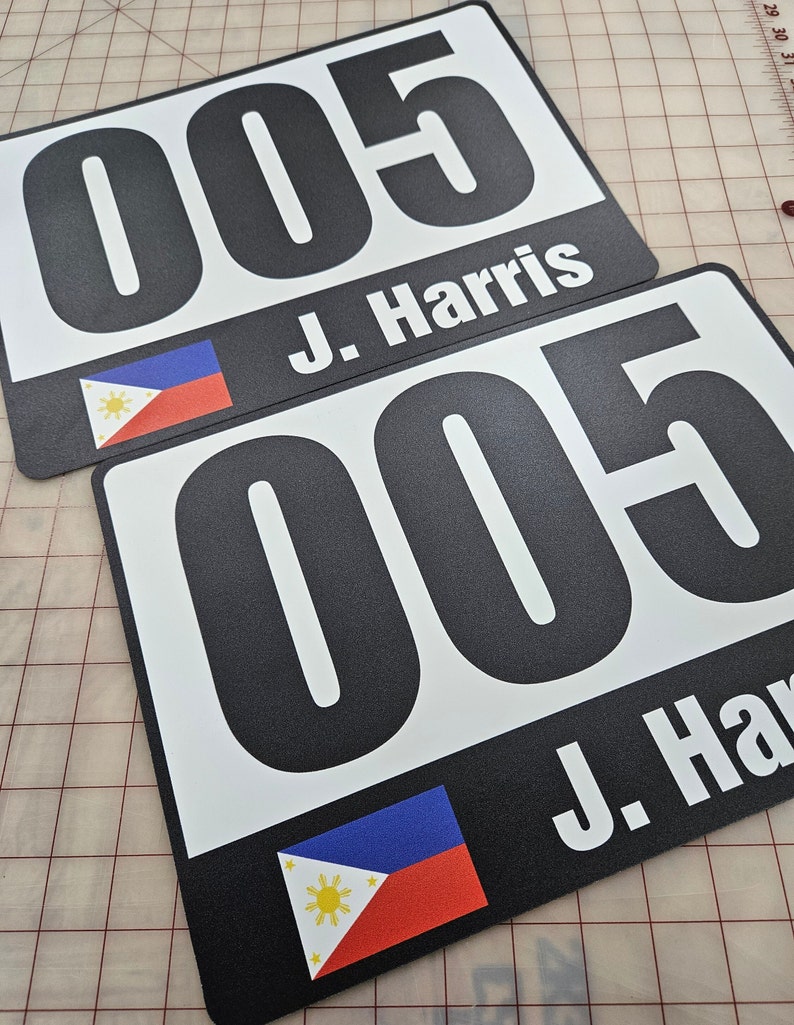 May include: Two rectangular race bibs featuring the large black number 005 printed on a white background. Below the number, the name J. Harris is printed in white text next to a small rectangular flag of the Philippines with blue, red, and white colors.