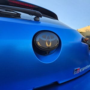 May include: A blue car with a black Toyota emblem on the rear. The emblem is surrounded by a chrome ring. The car also has a black "GR SPORT" emblem on the back.