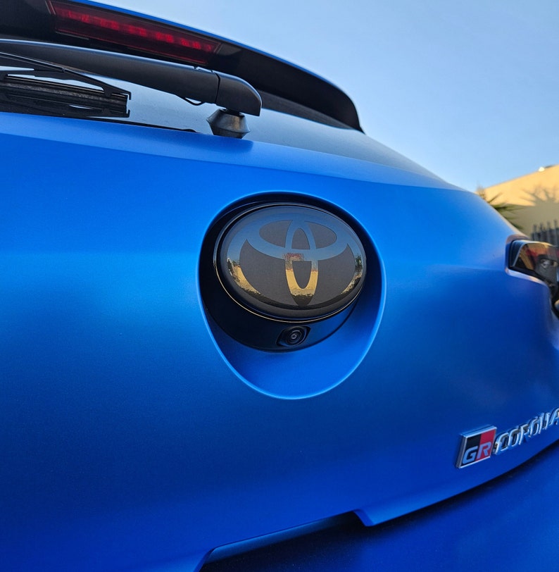 May include: A blue car with a black Toyota emblem on the rear. The emblem is surrounded by a chrome ring. The car also has a black "GR SPORT" emblem on the back.