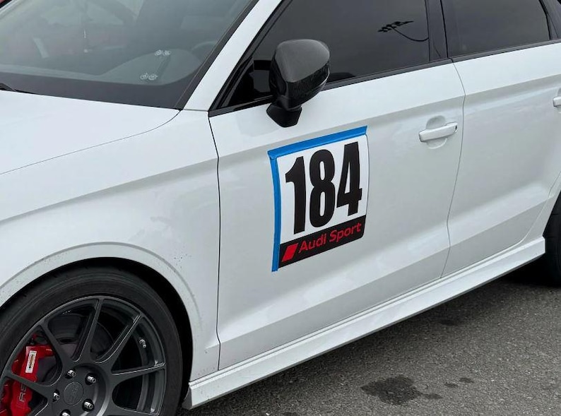 May include: A white Audi car featuring a black side mirror, dark grey wheels with red brake calipers, and a rectangular door decal displaying the number 184 in large black font above the red and white Audi Sport logo on a black background.
