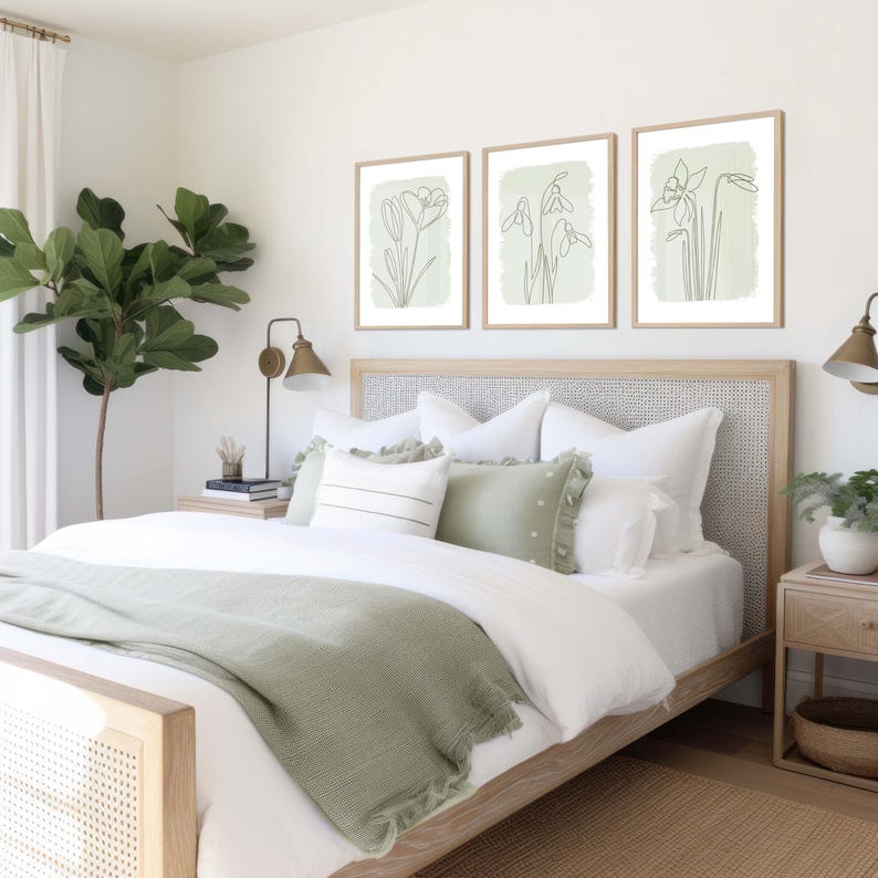 Op de afbeelding: Slaapkamer met een licht houten bed, wit beddengoed en een saliegroene sprei. Drie ingelijste botanische prints sieren de muur. Een grote potplant en twee nachtkastjes maken de inrichting compleet.