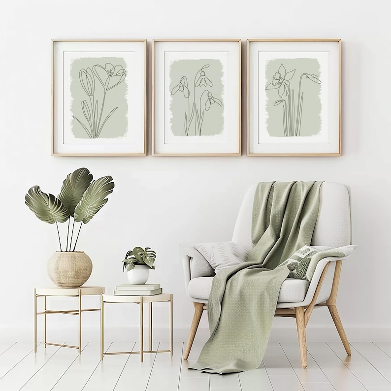 Op de afbeelding: Drie ingelijste botanische kunstprints met minimalistische lijntekeningen van bloemen in een lichtgroene tint. De frames zijn goudkleurig en de kunstwerken worden tentoongesteld boven een witte stoel met een groene deken. Een vaas met grote groene bladeren staat op een bijzettafel.