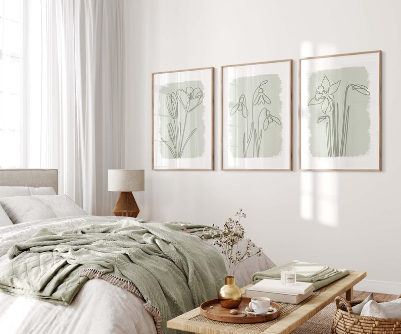 Op de afbeelding: Drie ingelijste botanische kunstprints met lijntekeningen van bloemen op een lichtgroene achtergrond. De prints zitten in houten frames en worden boven een bed met een groene deken getoond. De kamer heeft een neutraal kleurenpalet.