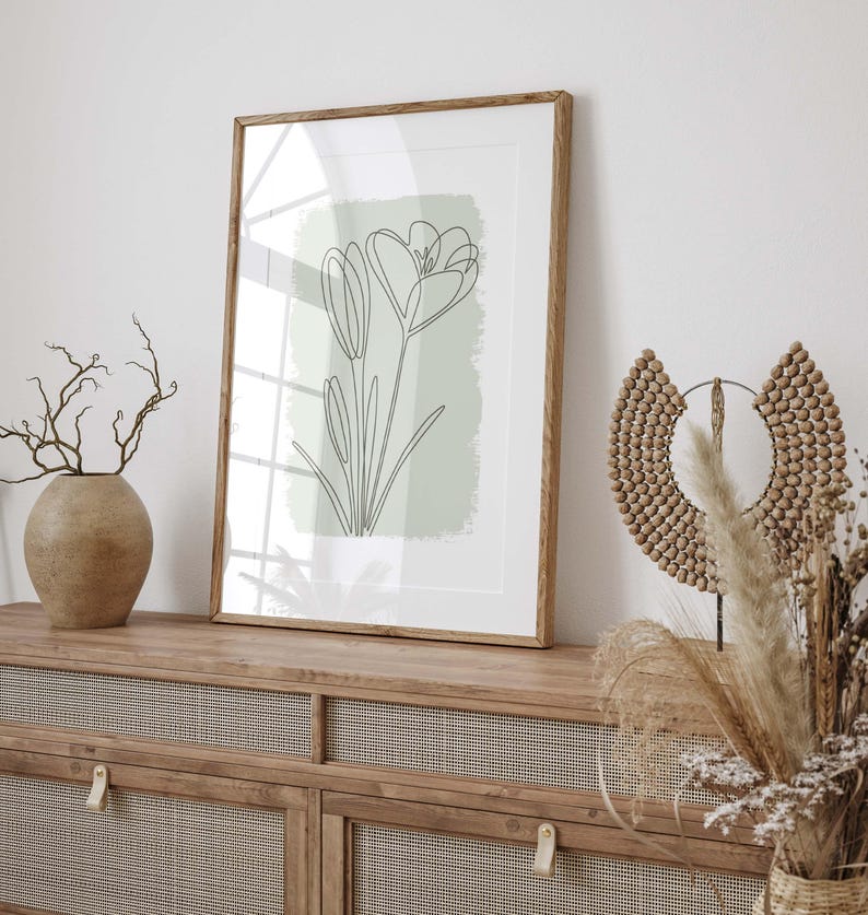 Op de afbeelding: Een ingelijste kunstdruk met een minimalistische lijntekening van een bloem op een lichtgroene achtergrond. Het kunstwerk zit in een houten lijst en wordt tentoongesteld op een houten kast. Decoratieve elementen zijn een vaas met takken en een kralen wandhanger.
