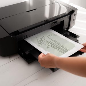 Op de afbeelding: Een zwarte inkjetprinter die een bloemenontwerp in lijnkunst afdrukt op wit papier. Het bloemenontwerp toont een narcis met lange stelen en bladeren. De printer staat op een wit houten oppervlak.