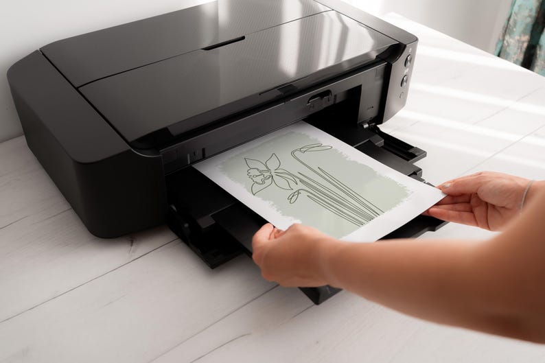 Op de afbeelding: Een zwarte inkjetprinter die een bloemenontwerp in lijnkunst afdrukt op wit papier. Het bloemenontwerp toont een narcis met lange stelen en bladeren. De printer staat op een wit houten oppervlak.