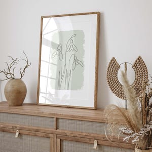 Op de afbeelding: Een ingelijste print met een minimalistische lijntekening van sneeuwklokjes op een lichtgroene achtergrond. Het kunstwerk wordt getoond op een houten kast, samen met decoratieve elementen zoals een vaas en een kralen wanddecoratie.