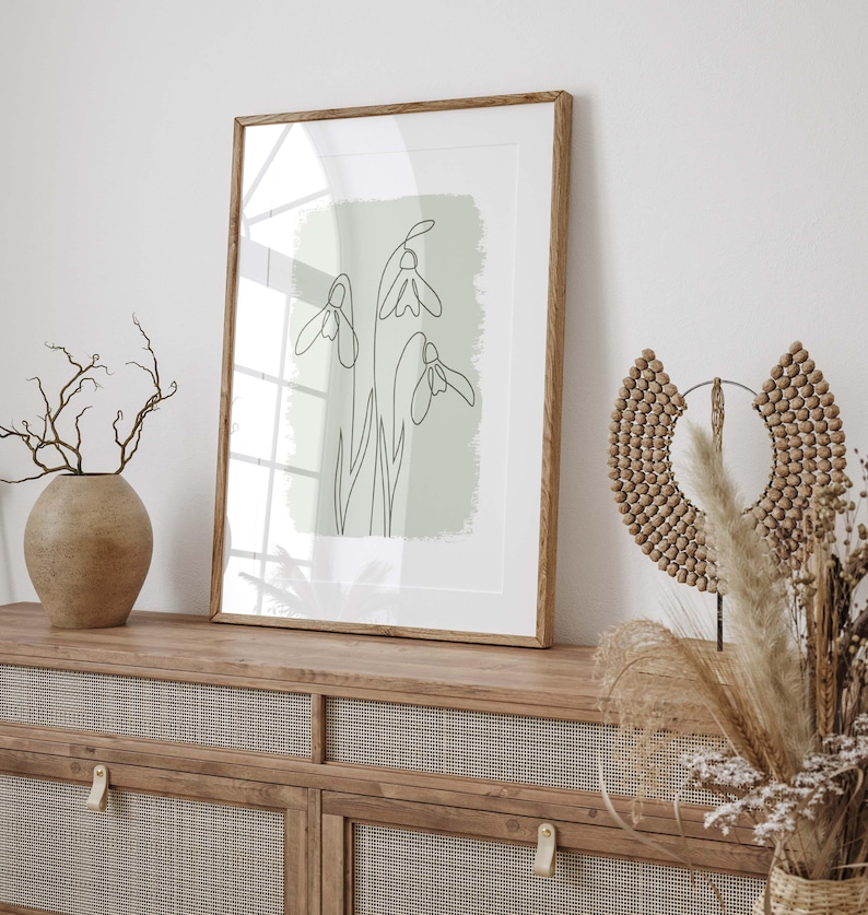 Op de afbeelding: Een ingelijste print met een minimalistische lijntekening van sneeuwklokjes op een lichtgroene achtergrond. Het kunstwerk wordt getoond op een houten kast, samen met decoratieve elementen zoals een vaas en een kralen wanddecoratie.