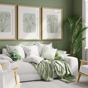 Op de afbeelding: Een woonkamer met een witte bank, stoelen en kussens. Drie ingelijste botanische kunstprints hangen aan de muur. Een groene plaid en kussens met bladerenpatronen zorgen voor kleur. Een potplant en een lamp zijn ook zichtbaar.