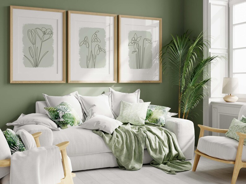 Op de afbeelding: Een woonkamer met een witte bank, stoelen en kussens. Drie ingelijste botanische kunstprints hangen aan de muur. Een groene plaid en kussens met bladerenpatronen zorgen voor kleur. Een potplant en een lamp zijn ook zichtbaar.