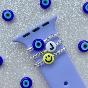 May include: Three silver beaded bracelets with charms on a purple Apple Watch band. The charms include a blue evil eye, a white letter J, and a yellow smiley face.