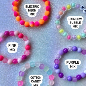 Puede incluir: Seis pulseras de cuentas coloridas con diferentes combinaciones de colores y texto. Las pulseras est&aacute;n hechas de cuentas peque&ntilde;as y redondas y tienen diferentes combinaciones de colores, incluyendo rosa, morado, azul, verde, amarillo y naranja. Las pulseras tienen texto en ellas, incluyendo "KIND", "ELECTRIC NEON MIX", "RAINBOW BUBBLE MIX", "PINK MIX", "COTTON CANDY MIX" y "PURPLE MIX".