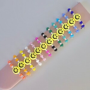 May include: A set of 12 colorful beaded bracelets with yellow smiley face charms. The bracelets are made with small, round beads in various colors, including blue, green, pink, orange, and white. Each bracelet features a single yellow smiley face charm.