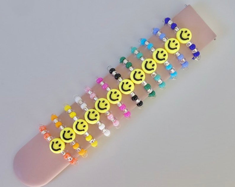 May include: A set of 12 colorful beaded bracelets with yellow smiley face charms. The bracelets are made with small, round beads in various colors, including blue, green, pink, orange, and white. Each bracelet features a single yellow smiley face charm.