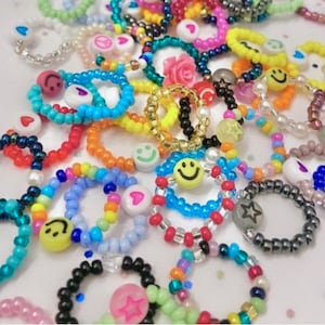 Puede incluir: Una colección de anillos de cuentas coloridas con varios encantos, incluyendo caras sonrientes, corazones, estrellas y una flor rosa. Los anillos están hechos de pequeñas cuentas coloridas y están apilados uno encima del otro.