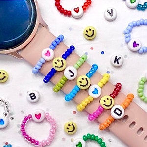 May include: A set of colourful beaded rings with smiley face, heart and letter charms. The rings are stacked on a pink wristband and a silver watch.