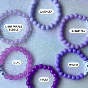 Puede incluir: Un conjunto de diez pulseras de cuentas coloridas en tonos de morado, lavanda y morado claro. Cada pulsera tiene un dije de letra que deletrea el nombre del color.