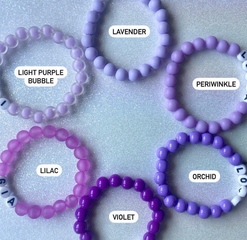 Puede incluir: Un conjunto de diez pulseras de cuentas coloridas en tonos de morado, lavanda y morado claro. Cada pulsera tiene un dije de letra que deletrea el nombre del color.