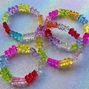 May include: Four colorful gummy bear bracelets in shades of red, pink, purple, yellow, green, blue, and orange. The bracelets are strung on a clear cord with silver beads. The background is a sparkly, iridescent surface.