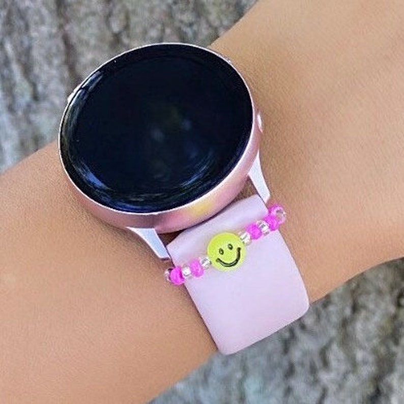 May include: A pink smartwatch with a black face and a pink and clear beaded bracelet with a yellow smiley face charm.