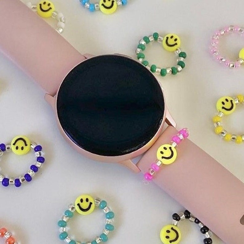 May include: A pink smartwatch with a black face and a variety of colorful beaded bracelets with smiley face charms. The bracelets are made with different colored beads, including pink, green, blue, yellow, and black.
