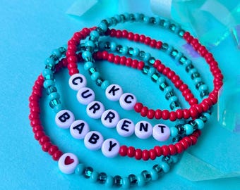 May include: A set of four beaded bracelets with the words "BABY", "CURRENT", "KC", and a heart charm. The bracelets are made with red and turquoise beads.