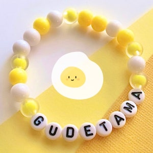 May include: A yellow and white beaded bracelet with the word "Gudetama" spelled out in black letters. The bracelet is on a yellow surface with a cartoon image of a fried egg.