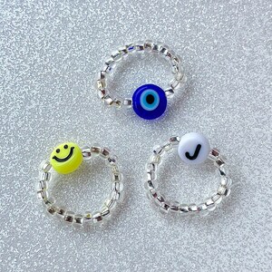 May include: Three silver beaded rings with different charms. The first ring has a blue evil eye charm, the second ring has a yellow smiley face charm, and the third ring has a white charm with a black checkmark.